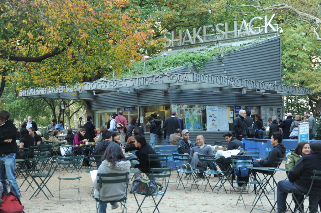 Photograph of Shake Shack. The tables and chairs in front of the structure are almost full with people.