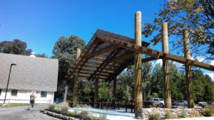 A photograph of the Hunt House structure, seen adjacent to a parking lot and with picnic tables underneath. The structure features 6 wood poles (3 on either side), wood slats forming a pitched roof, and steel cables and wood beams reinforcing the structure.