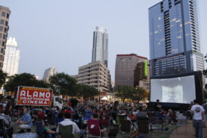 A crowd is gathered on lawn chairs and blankets to watch a movie on an inflatable screen at the park.