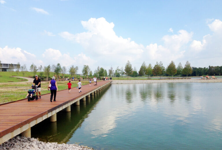 Image of a boardwalk at Shelby Farms at the edge of the lake. Visitors are seen walking and running across the boardwalk. Trees and the playground can be seen in the background.