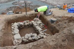 An archeaologist is excavating a historical privy. The privy is made of several layers of stone stacked to form the outline of a square.