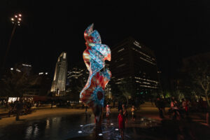 A colorful, tall, narrow sculpture in a shallow pool of water is illuminated by uplights at night. A few children gathered around in the water.