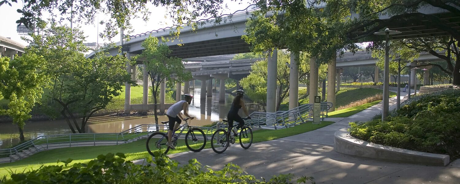 Two bicyclers biking on a park path that goes under roadway bridges. The bayou can be seen in the background.