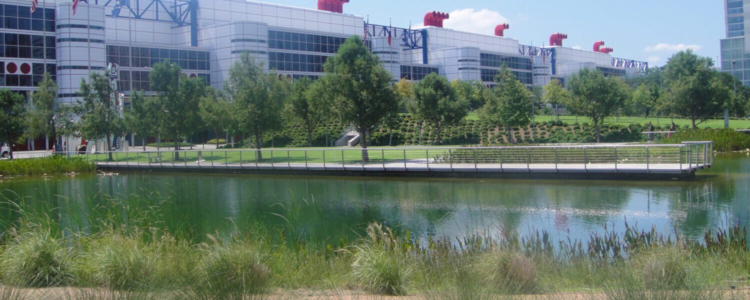 A cropped photograph where grass plants border the lake at Discovery Green. An overlook, gardens, and the Houston Convention Center are in the background.