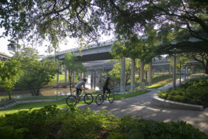 Two bicyclers biking on a park path that goes under roadway bridges. The bayou can be seen in the background.