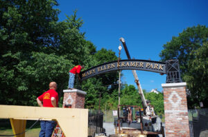 A photograph of the installation of the park entry sign. A small crane is hoisting the sign between two columns while a man works to attach it to one column.