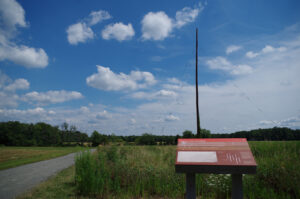 Photograph of the "last pole standing" at the park and its interpretive sign. The tallness of the pole is striking against the blue sky. It is one of 4 rhombic antenna poles erected in 1940 that enabled wireless overseas telephone calls to Tel Aviv, Israel.