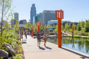 Groups of people walk along a wide path next to a rectangular pool. Sculptural orange pole lights line the border of the pool.